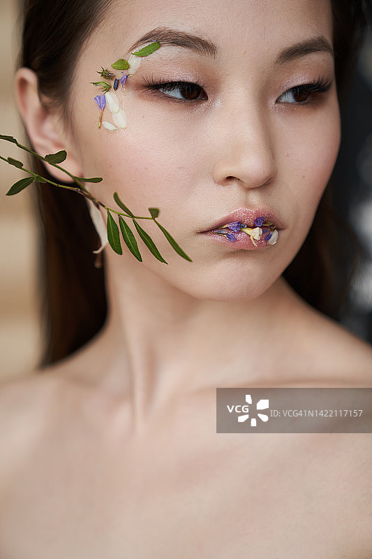 美丽的亚洲女人与黑色的头发和时尚的妆容看远离。小花瓣粘在年轻女子的嘴唇和眼睛周围。一片绿叶贴在模特的脸颊上。米色背景上的天然化妆品概念。图片素材