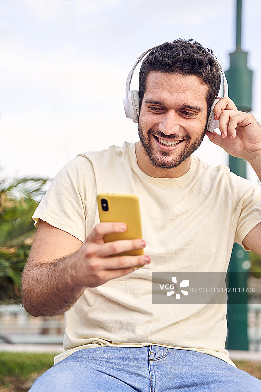 在城市的街道上，一个穿着休闲装的年轻男子用无线耳机和手机听音乐。图片素材