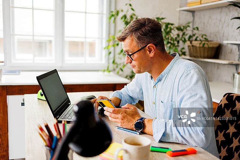 一名男子在家庭办公室使用笔记本电脑和智能手机在线学习图片素材