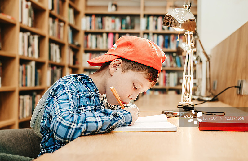 一个戴红帽子的男孩坐在图书馆的桌子上在笔记本上写着什么。准备作业。图片素材