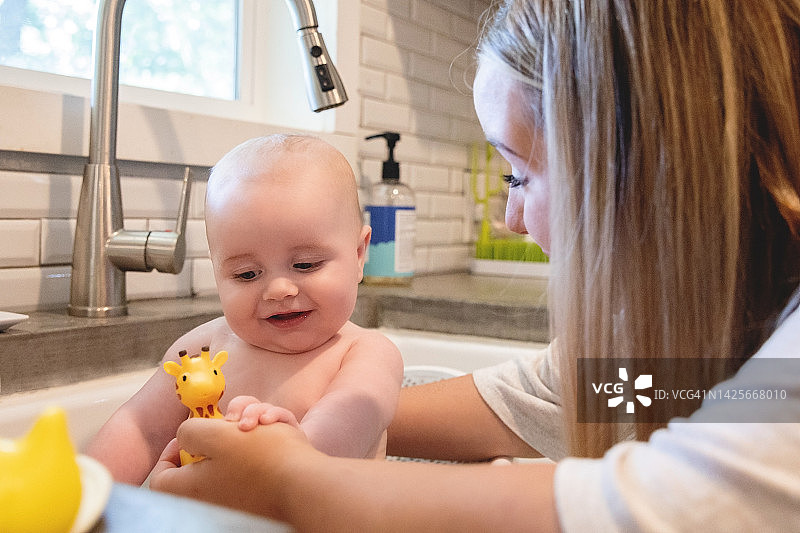 一位年轻的母亲正在家里的厨房水池里给襁褓中的儿子洗澡，并用毛巾把他裹起来图片素材