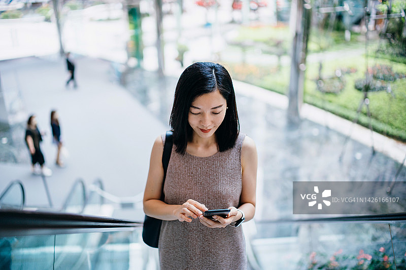 年轻的亚洲女商人在办公大楼上班，乘坐自动扶梯时使用智能手机。忙碌的生意图片素材