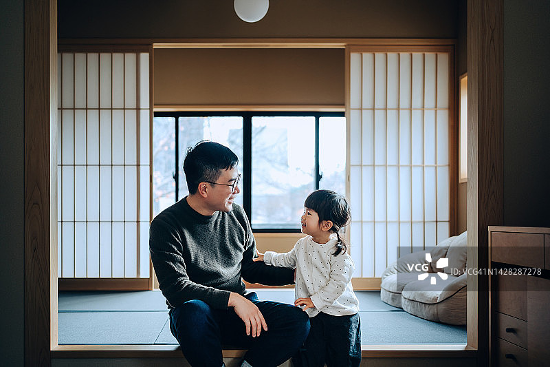 年轻可爱的亚洲父亲和她可爱的小女儿坐在传统日式木制公寓的榻榻米上，聊天，享受父女亲密的时光。家庭生活方式。爱与关怀概念图片素材