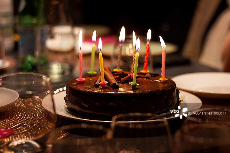 节日餐桌上的巧克力生日蛋糕画像，上面点着蜡烛。点燃的蜡烛都有不同的颜色。图片素材