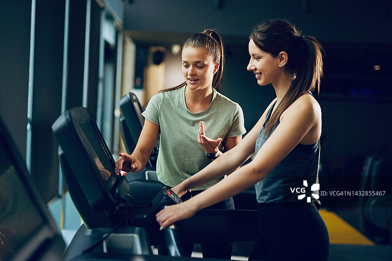 女教练协助年轻女子在健身房的跑步机上锻炼图片素材