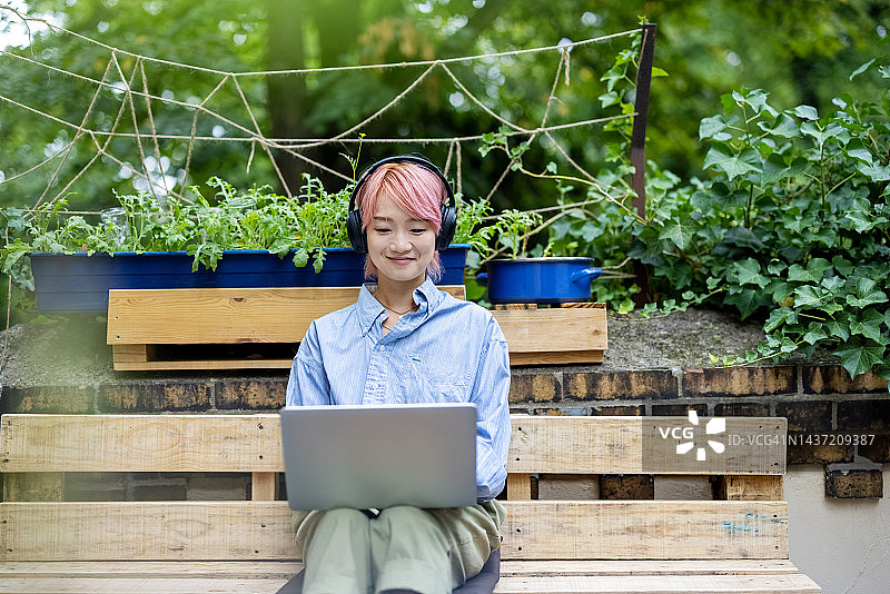 戴着耳机的亚洲妇女坐在办公室外的长凳上用笔记本电脑工作图片素材
