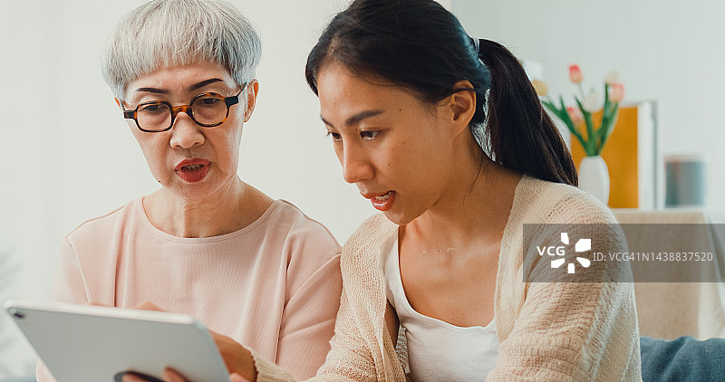 幸福的亚洲家庭，年幼的女儿和年长的妈妈教年长的妈妈使用平板电脑，在家里指着屏幕，向孩子询问有关移动设备的问题。图片素材
