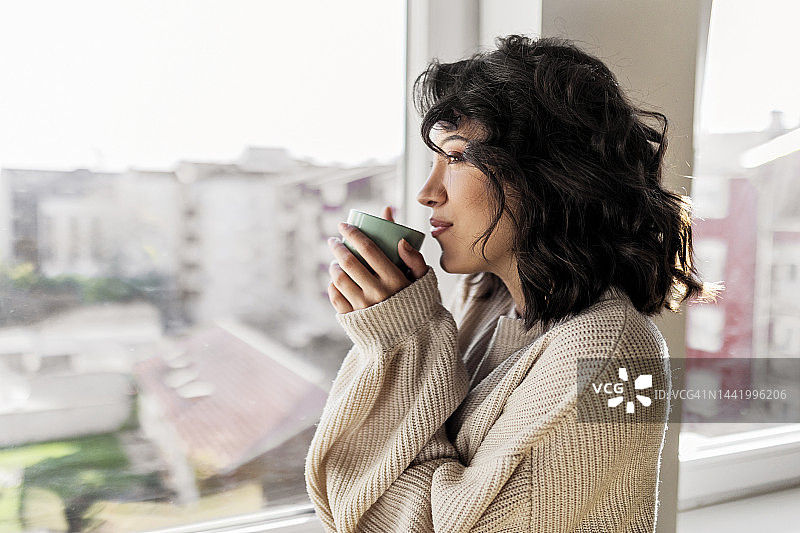 年轻女子一边喝着咖啡，一边透过窗户往外看图片素材