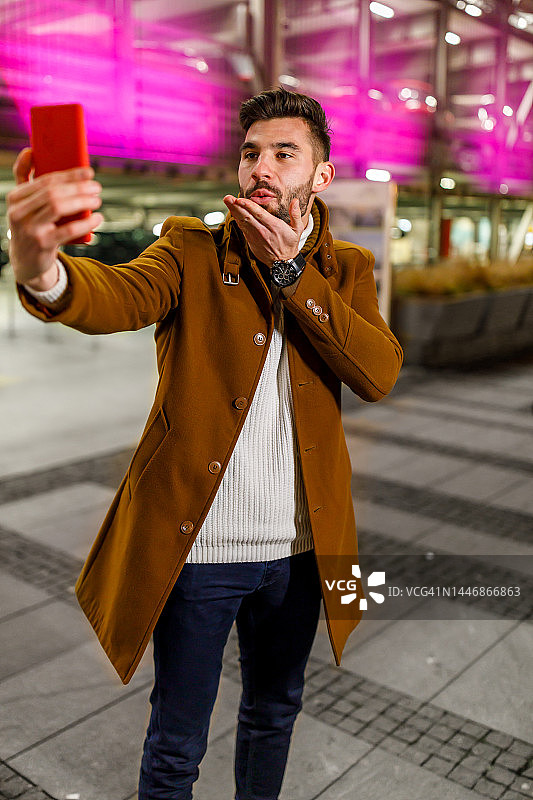 年轻男子在夜晚的城市街道上用手机自拍。图片素材