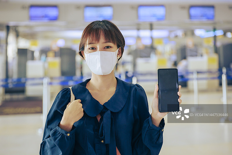 年轻的亚洲女商人戴着口罩，手持空屏手机在机场离境区。商务旅行的概念。图片素材