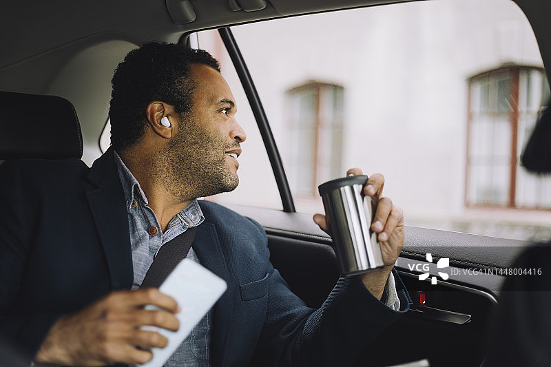 微笑的男性通勤者，带着隔热的饮料容器和智能手机从车窗向外看图片素材