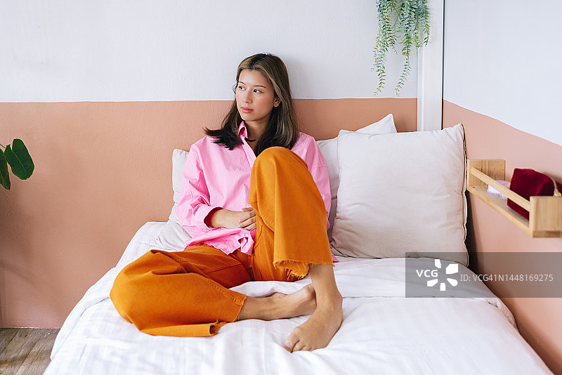 一个穿着粉红色衬衫和橙色裤子的快乐美丽的女人在家里放松时看着别处图片素材
