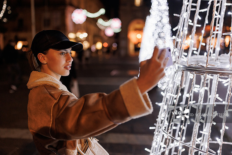夜晚，微笑的年轻女子在圣诞装饰的城市用手机自拍图片素材
