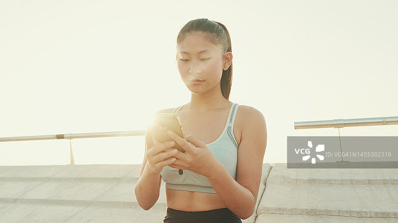 在晨光中，站在现代建筑背景的海滨，微笑的亚洲女孩在运动上衣使用手机图片素材