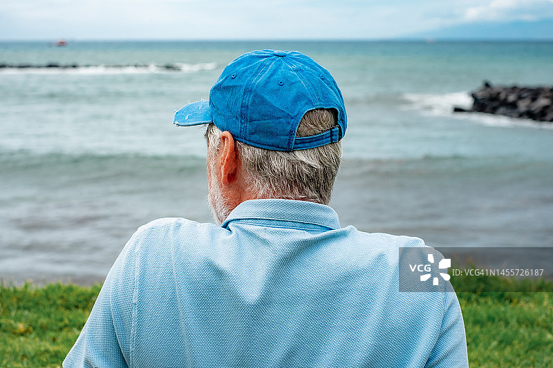 在冬天的一天，高级男子坐在海滩上享受空闲假期或退休的背影。大胡子放松的人戴着蓝色的帽子看着海面上的地平线图片素材