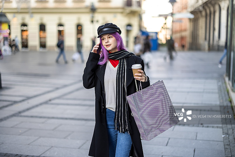 一位美丽的年轻女子走在城市的街道上，喝着咖啡，用着电话。图片素材