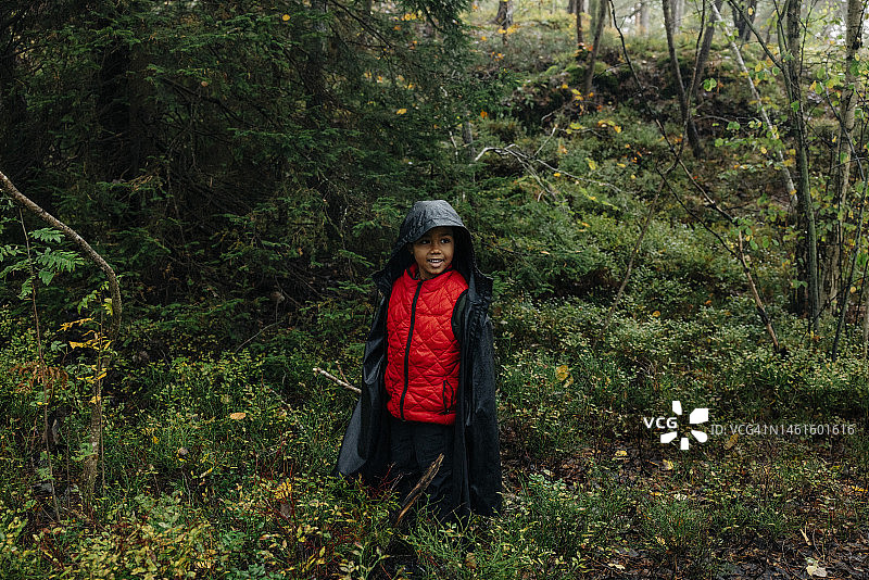 穿着雨衣的微笑男孩站在森林里的植物中看着别处图片素材