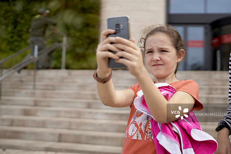 在宽阔的楼梯上，一个棕色头发，白色皮肤，穿着便服的女孩双手拿着手机，背景中有一个人有点模糊。图片素材