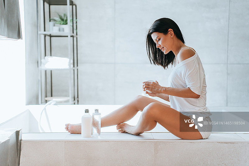 黑发西班牙年轻女子坐在浴室应用乳液在皮肤上。美丽的白人女性在护肤过程中使用乳霜腿部。医疗保健，早上的女人例行公事身体护理，保湿图片素材