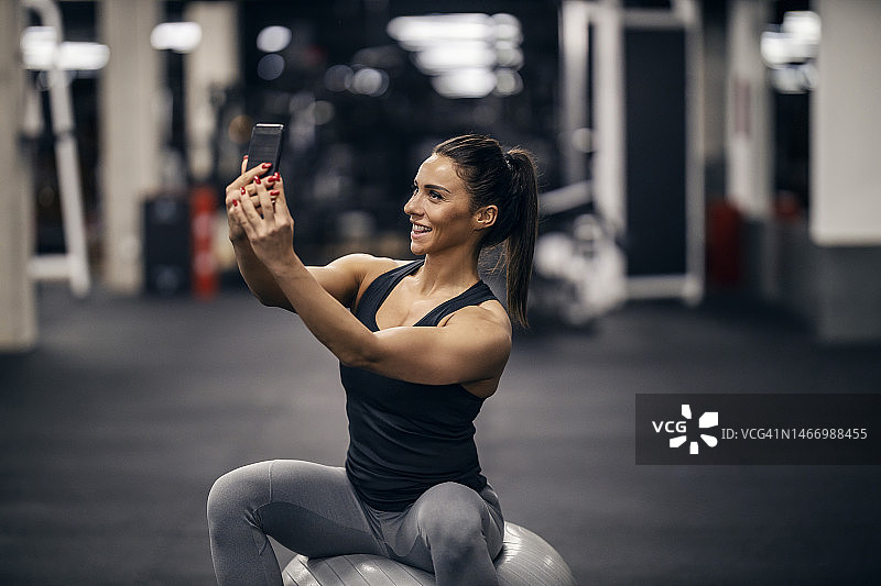 一位快乐的健身女运动员坐在健身房的普拉提球上自拍。图片素材