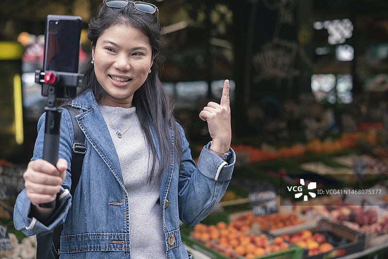亚洲女性博主在城市的露天市场用自拍杆拿着手机录制视频。年轻的亚洲女游客女子手持智能手机，录制视频旅游概念图片素材