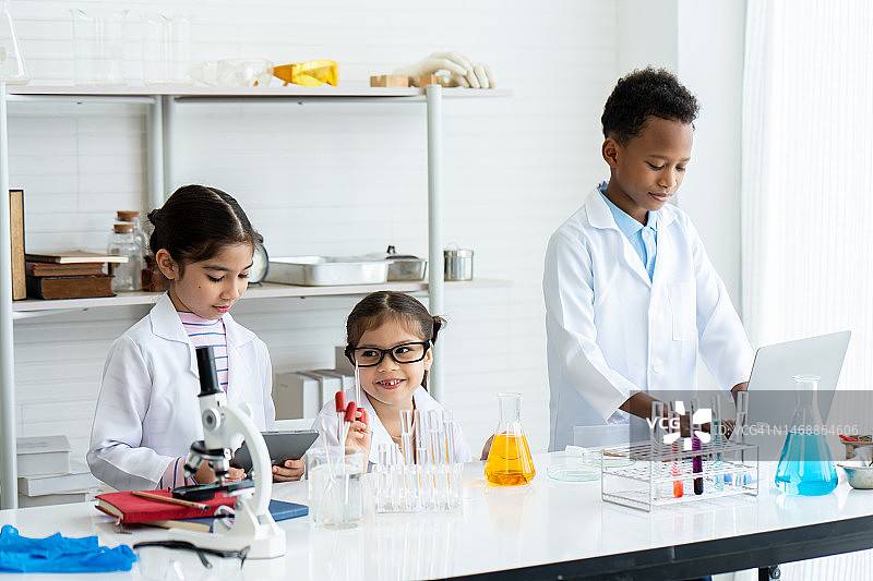 两个亚洲小女孩和一个穿着白大褂的非洲小男孩，架子和桌子上放着许多实验工具。在实验课上互相帮助，检查并记录结果信息。图片素材