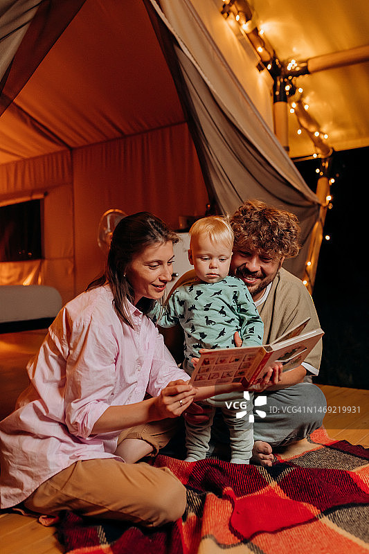 快乐的家庭与可爱的宝宝一起玩耍和阅读在夏季晚上豪华露营在舒适的篝火旁。户外休闲休闲的豪华露营帐篷。生活方式的概念图片素材