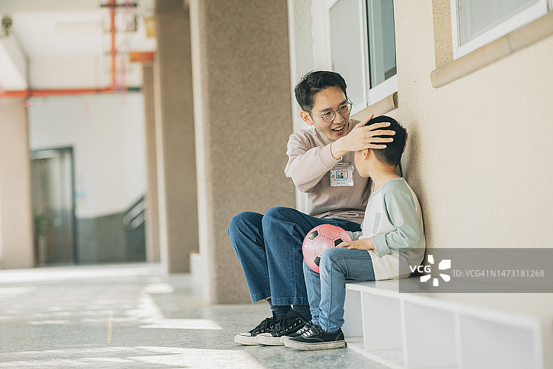 一位亚裔幼儿园老师坐在教室外面，正在和一个灰心丧气的孩子交谈。她全神贯注地倾听，安慰孩子。图片素材