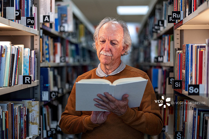 一位年长的白人男子在图书馆的书架间阅读一本白色的书图片素材