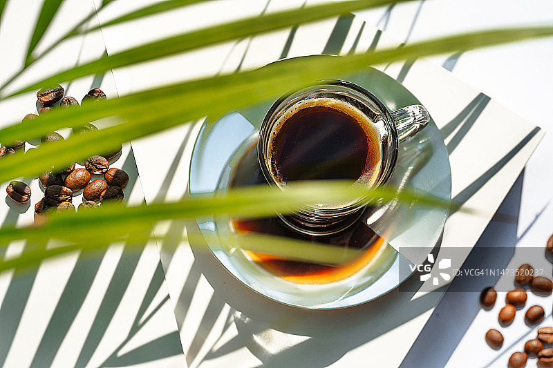 芳香的早晨咖啡在玻璃杯和烘焙咖啡豆与绿色的棕榈树叶子的阴影在白色的背景。图片素材