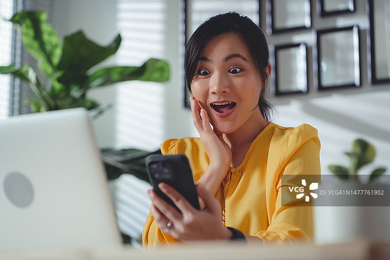 亚洲女性坐在家里办公室用智能手机，开心兴奋。图片素材