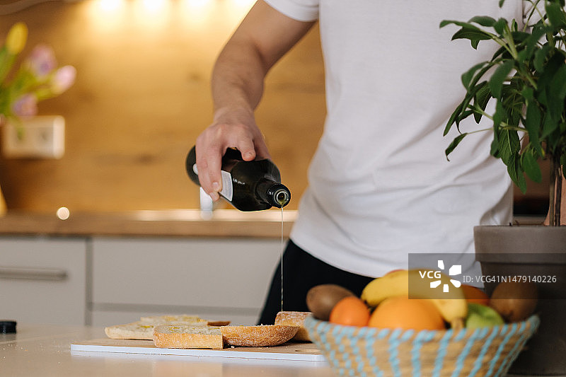 男子在长棍面包上涂橄榄油的特写镜头。明亮的厨房内部图片素材