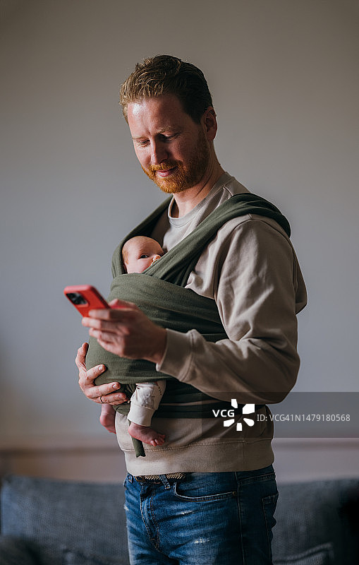 一个快乐的姜色男人一边用手机一边照顾看着镜头的可爱的新生儿图片素材