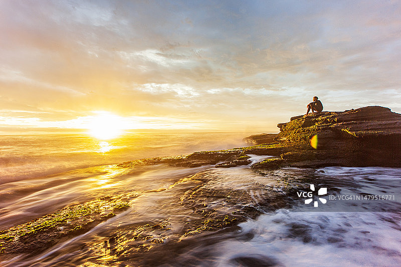 长时间曝光的海景水流过岩石与年轻人观察金色的日落图片素材