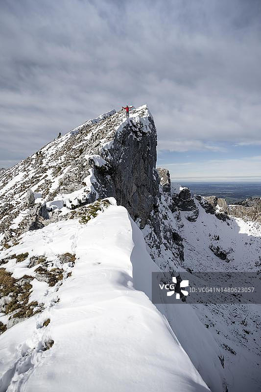 身穿红色夹克的登山运动员站在岩石塔尖上，冬天有雪的山脉，徒步前往德国巴伐利亚阿默高阿尔卑斯山的阿默高Hochplatte图片素材