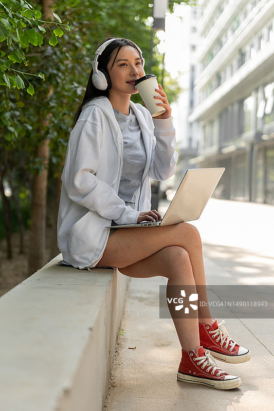 戴着耳机的年轻微笑女子在笔记本电脑上工作，坐在户外喝着外卖咖啡图片素材