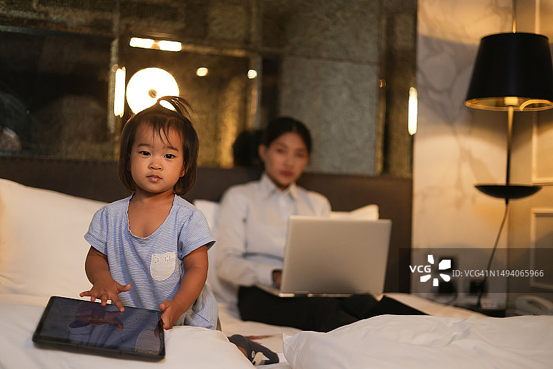 单身年轻的亚洲母亲在笔记本电脑上打字，在网上工作，而她的孩子在她旁边的床上玩耍图片素材