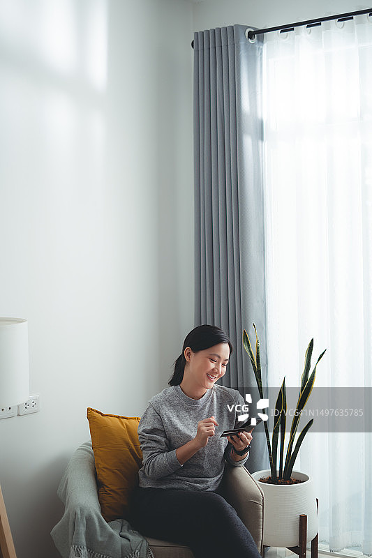 亚洲女性坐在扶手椅上开心地微笑着，用智能手机和信用卡在家里网上购物。图片素材