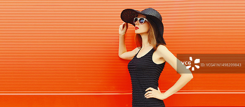 美丽的年轻女子的肖像，戴着黑色的夏季草帽，穿着裙子，在橙色的背景上看向别处，广告文字的空白复制空间图片素材