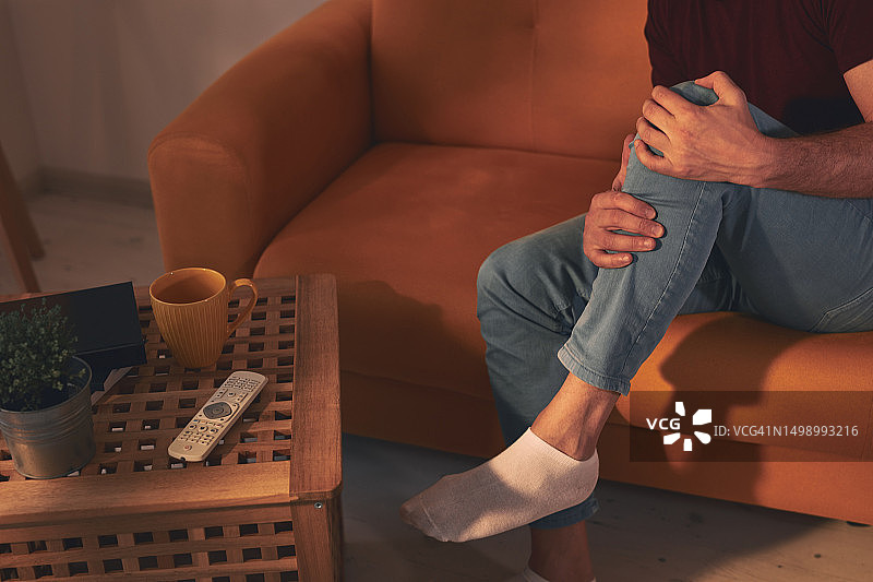 一个膝盖疼痛的男人坐在家里的沙发上。图片素材