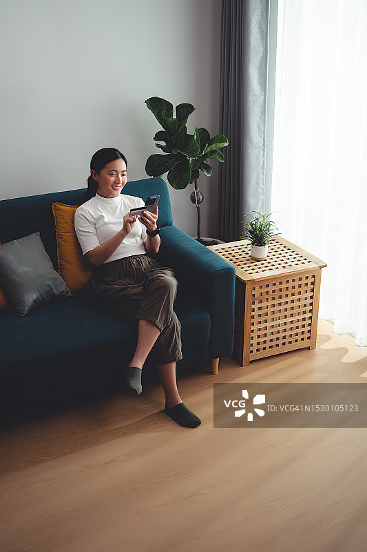 亚洲女性开心兴奋地坐在沙发上用智能手机和信用卡在家网上购物。图片素材