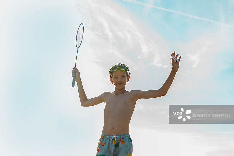 多云的蓝天下，一个面带微笑的少年拿着羽毛球拍和羽毛球图片素材