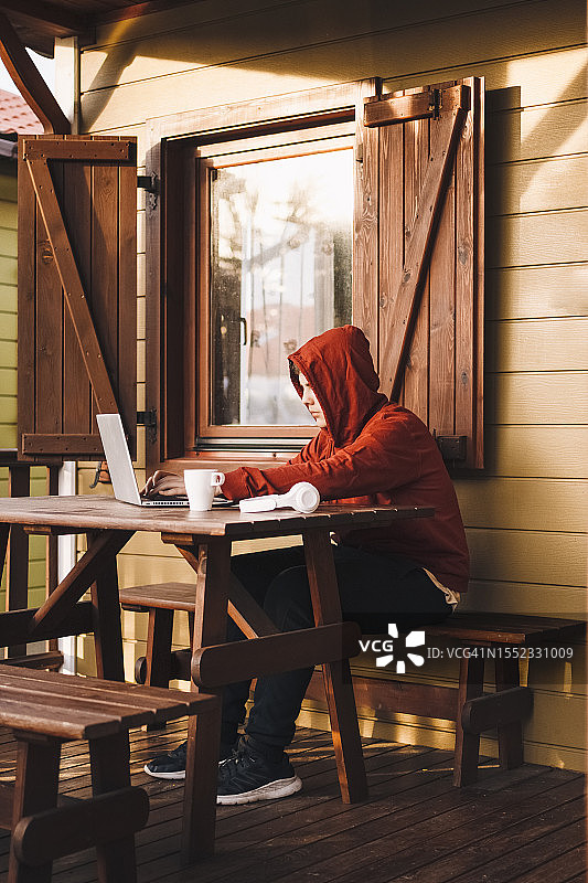 穿着红色连帽毛衣的少年在小木屋的门廊上使用他的笔记本电脑。图片素材