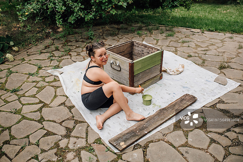 可爱的小女孩坐在铺好的后院塑料布上，把旧木箱漆成新鲜的绿色图片素材
