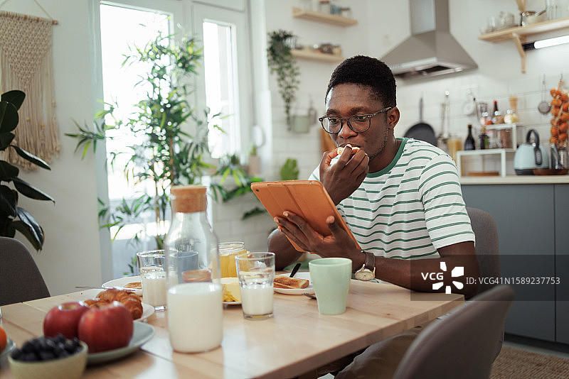 一个年轻人在家庭厨房吃早餐时使用平板电脑图片素材