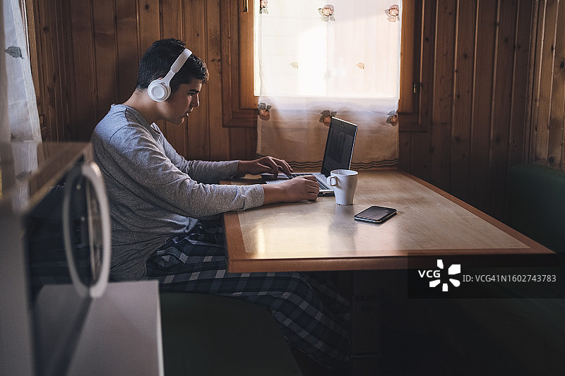 一个穿着睡衣，戴着无线耳机的少年，一大早坐在小木屋里的一张桌子上，喝着咖啡，用着笔记本电脑。图片素材
