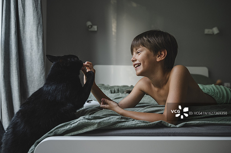 微笑的男孩躺在床上和家里的黑猫玩耍图片素材