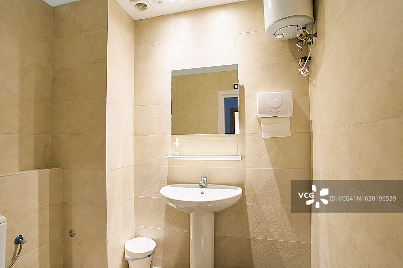 浴室的一部分，展示了一个水槽、一面镜子、纸巾、一个热水器、一个塑料废纸篓和一面贴着米色瓷砖的墙。图片素材