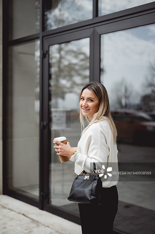 一个微笑的金发女人端着咖啡走在街上。图片素材