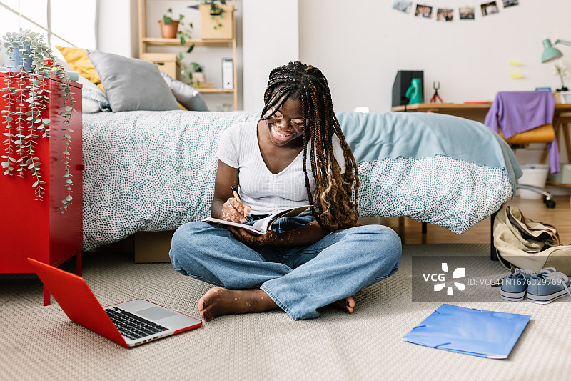 高中女生在家里一边学习一边记笔记。年轻的黑人女性坐在卧室地板上用笔记本电脑上网学习。教育与科技理念。图片素材